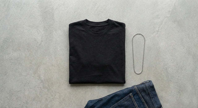 A folded black cotton t-shirt flat lay with dark denim jeans and a silver chain on a concrete surface.