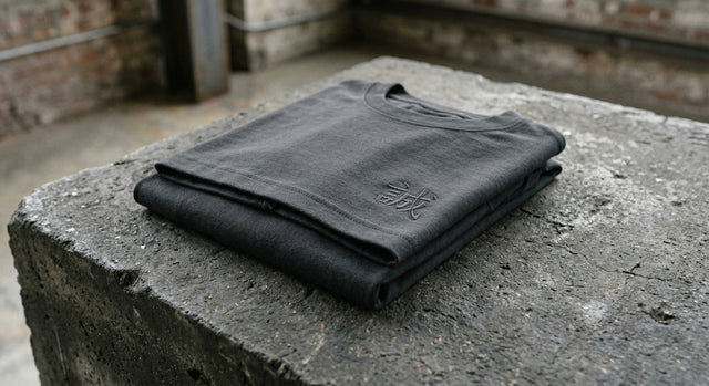A close-up shot of a charcoal grey folded t-shirt with subtle Kanji embroidery on concrete.