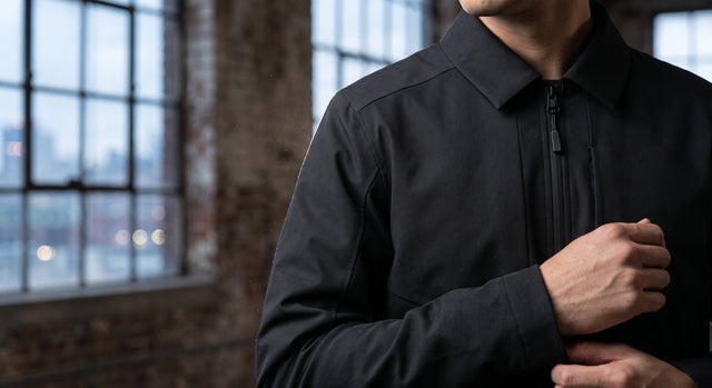 Close-up of a structured, matte black commuter jacket against a blurred Brooklyn warehouse window at dusk.