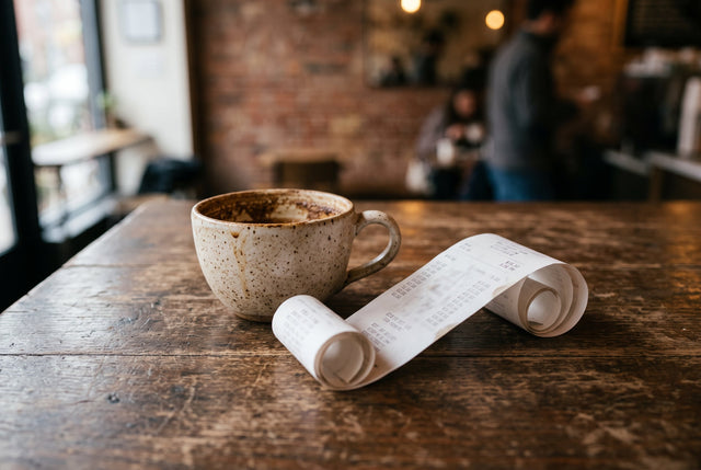 A curled receipt lies beside a coffee cup on a worn cafe table in soft afternoon light.