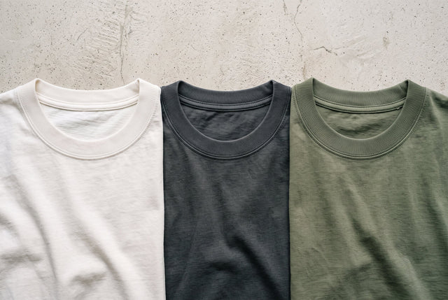 Three cotton tees with different crew neck openings arranged on a pale concrete surface.