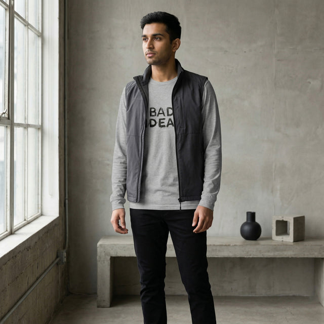 Man wearing a gray jacket and black pants standing in a room with concrete walls and a window.