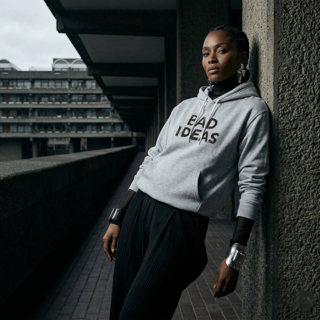 Person wearing a hoodie with 'BAD IDEAS' text, leaning against a wall in an urban setting.