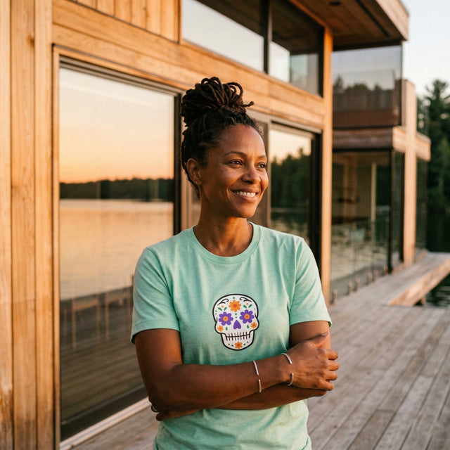 Woman wearing a light blue t-shirt with a colorful skull design, standing in front of a wooden cabin by a lake.