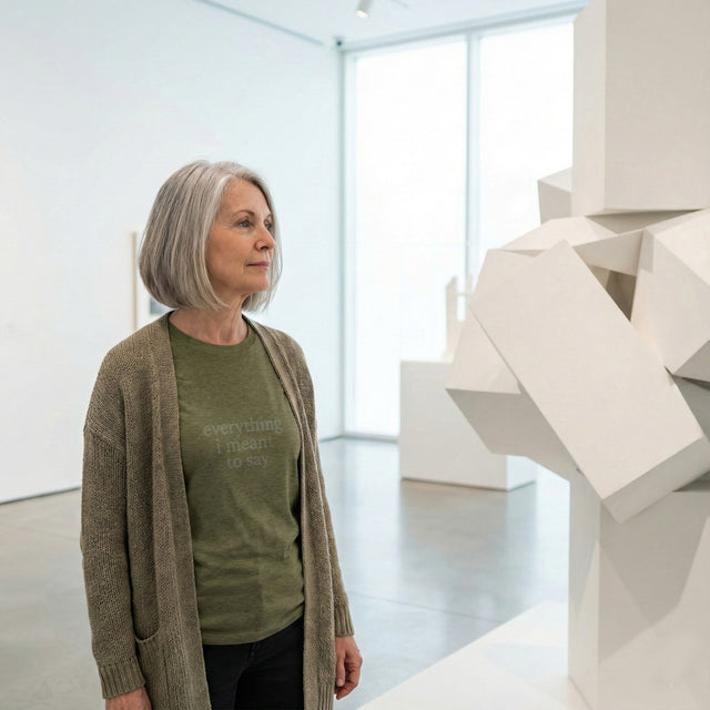 Heather olive green t-shirt featuring the minimalist grey text "everything i meant to say", worn by a mature woman standing in a quiet modern art gallery.