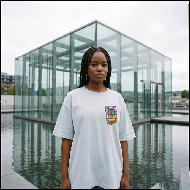 A young woman stands in front of a modern glass pavilion with water in the foreground, wearing an oversized white t-shirt with a colorful graphic on the left chest.