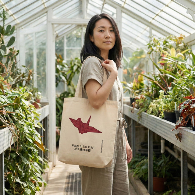 Ichinichi "Peace In The Fold" beige canvas tote bag with red origami crane graphic, held by a Japanese woman in a botanical greenhouse setting.