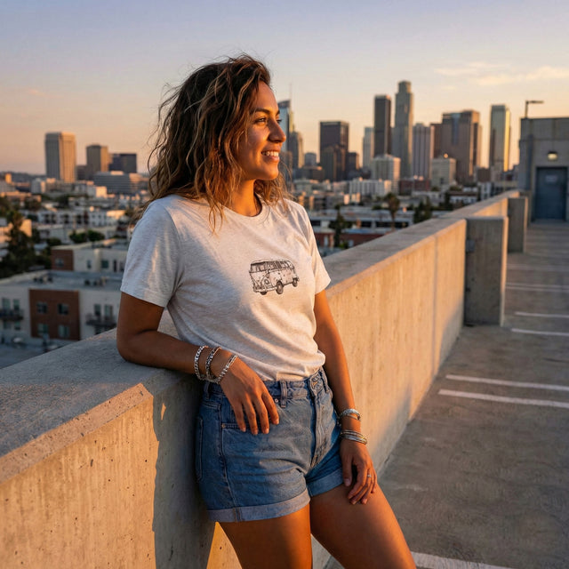 Hispanic woman wearing white t-shirt with vintage VW bus sketch, posing on a city rooftop parking garage at sunset.