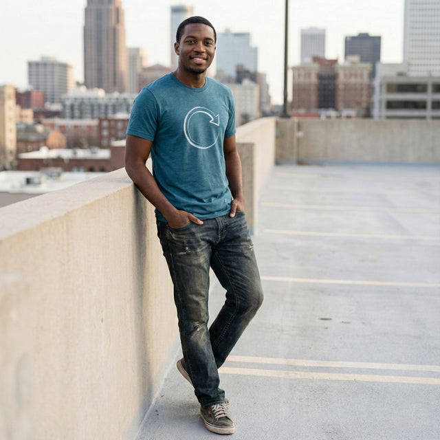 Young Black man modeling a heather deep teal t-shirt with a white brushstroke refresh loop icon, standing on an urban rooftop parking deck with a blurred city skyline in the background.