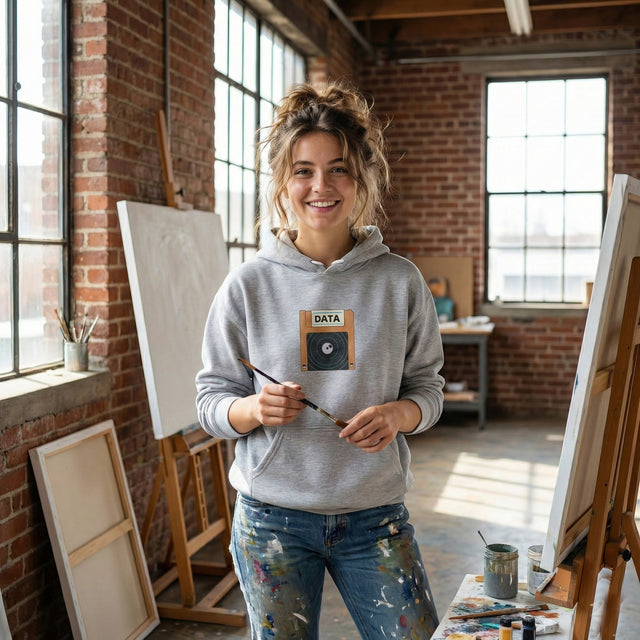Woman in a gray hoodie with a graphic design, holding a paintbrush in an art studio.
