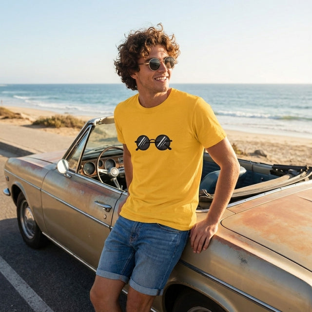 A young man with curly hair and round sunglasses leans against a vintage car on a sunny coastal road, wearing a bright yellow t-shirt with a sunglasses graphic.