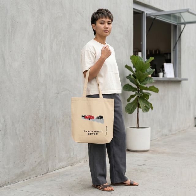 Person holding a beige tote bag with a graphic design, standing outdoors against a light gray wall.