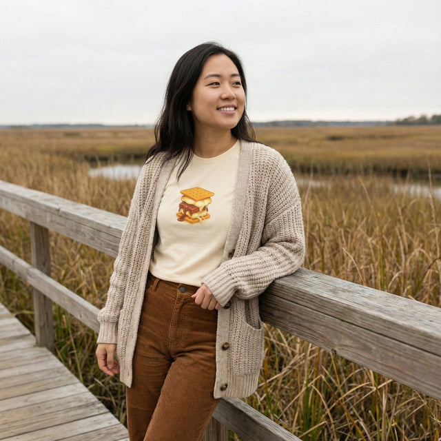 Woman standing on a wooden bridge in a natural setting wearing a beige cardigan and brown pants.