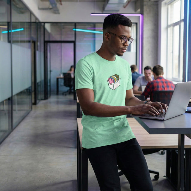Ichinichi Unisex Staple T-Shirt in Heather Mint featuring a graphic of a white storage bin filled with colorful folders, worn by a male model in a modern co-working office space.