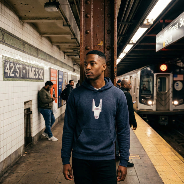 Man in a subway station wearing a blue hoodie with a logo, 42nd Street Times Square station visible.
