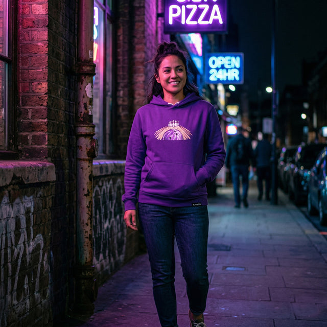 Person wearing a purple hoodie with a logo on a neon-lit street at night.