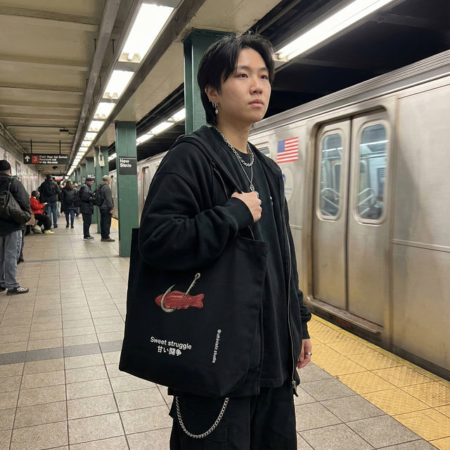 Black canvas tote bag featuring a red Swedish fish candy on a hook graphic and "Sweet struggle" text, carried by a model on a busy city subway platform.