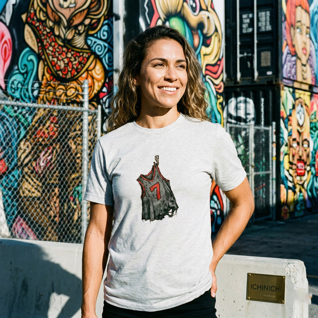 A sharp lifestyle photograph of a 30s Brazilian woman smiling, standing at Wynwood Walls in Miami. She is wearing the 'Echoes of Effort' grey t-shirt featuring the tattered basketball jersey graphic. Hard, direct sunlight creates defined shadows and highlights the textures of the shirt and the colorful graffiti murals in the background.