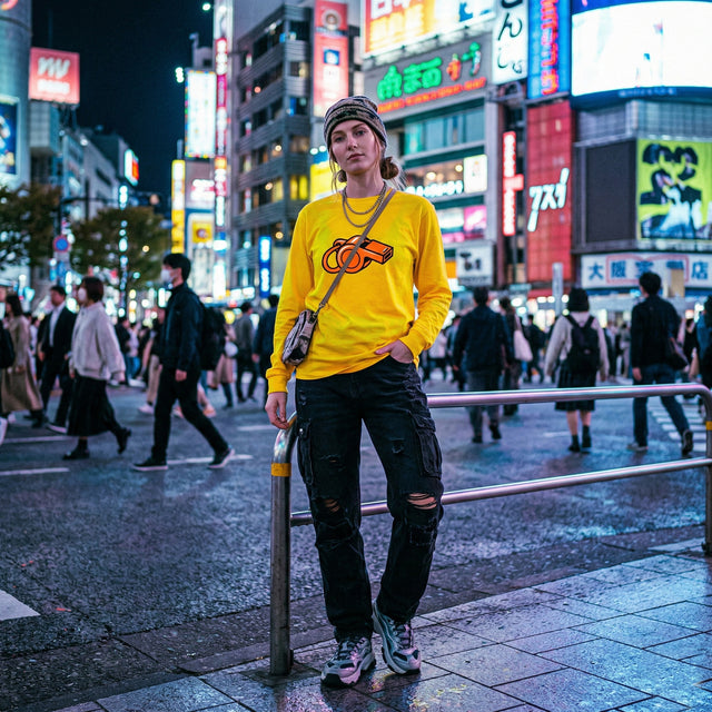 A young Caucasian woman with a streetwear aesthetic wearing a bright gold long-sleeve tee featuring an orange whistle graphic, standing amidst the vibrant neon lights of Shibuya at night.