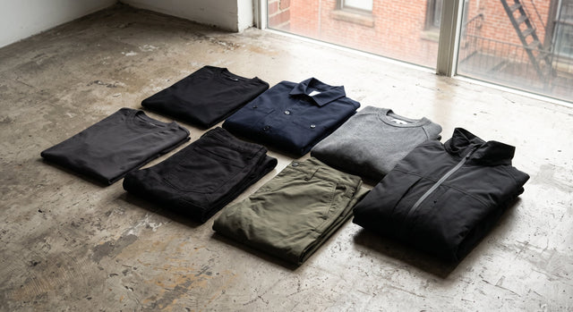 Editorial flat lay of 7 minimalist streetwear items on a raw concrete floor.