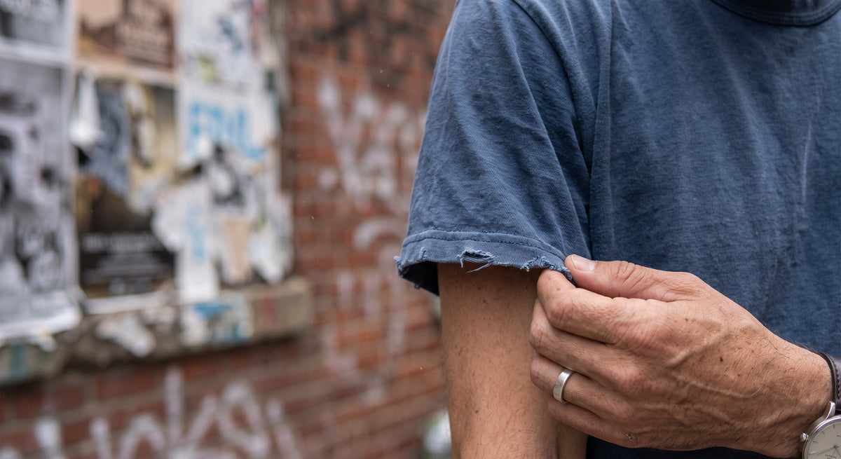 Extreme close-up of a hand gently touching the frayed cuff of a faded indigo heavy cotton t-shirt, Brooklyn background.