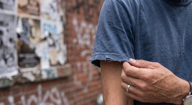 Extreme close-up of a hand gently touching the frayed cuff of a faded indigo heavy cotton t-shirt, Brooklyn background.