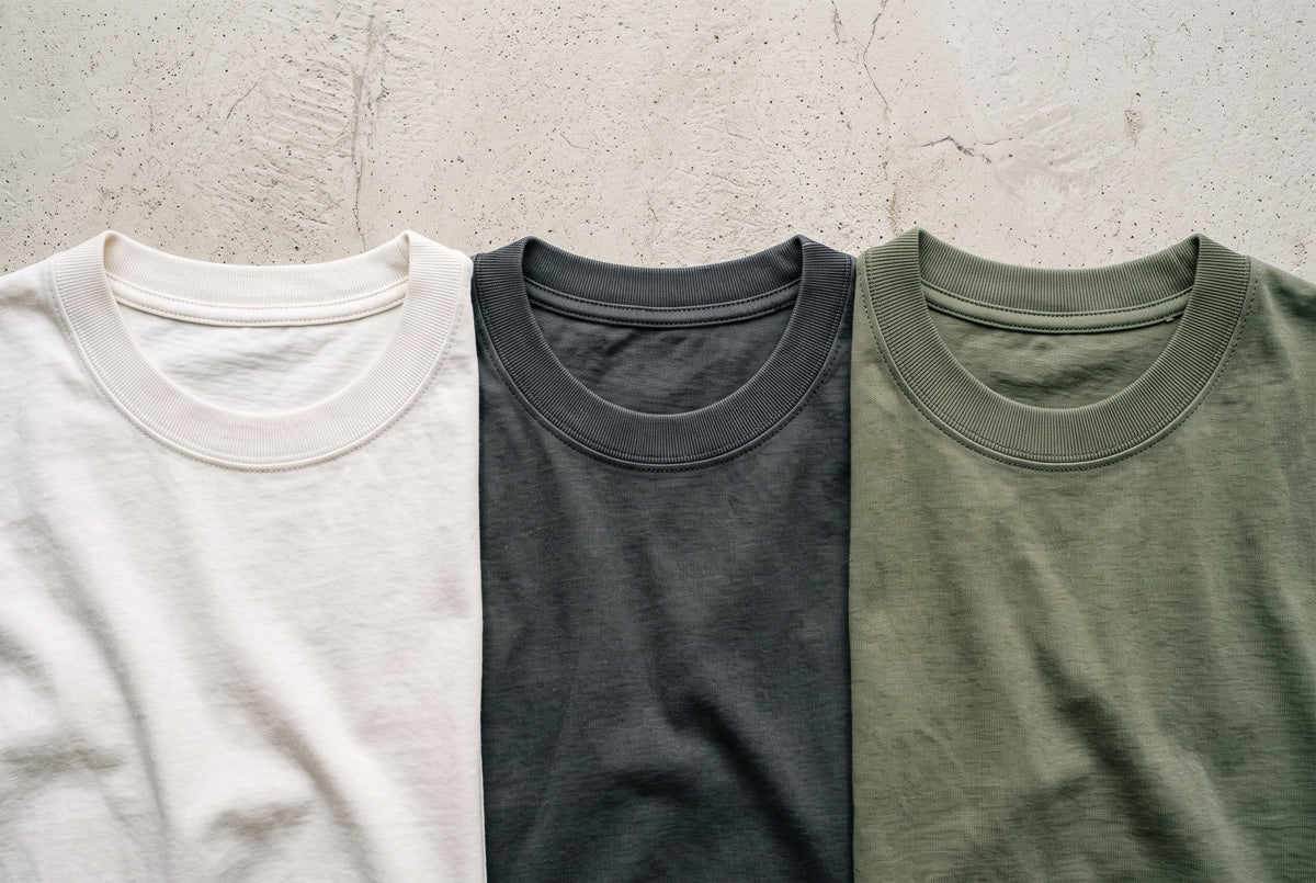 Three cotton tees with different crew neck openings arranged on a pale concrete surface.