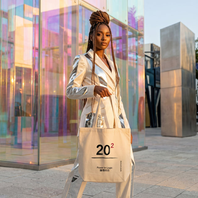 Woman holding a beige tote bag with '202' design, standing in front of modern glass architecture.