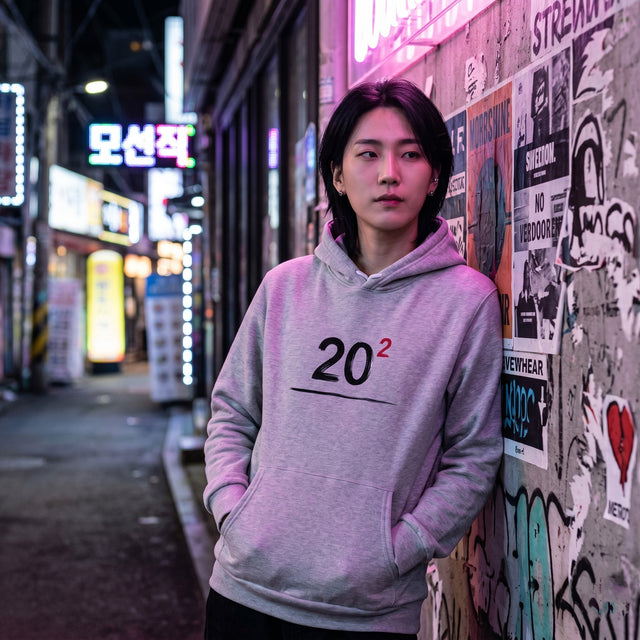Person wearing a gray hoodie with '20²' design, standing against a graffiti-covered wall in an urban setting.