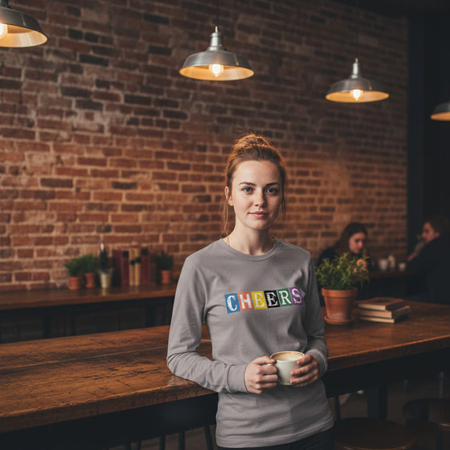 Woman in a gray long sleeve shirt with 'CHEERS' in colorful letters, holding a cup in a cozy indoor setting.