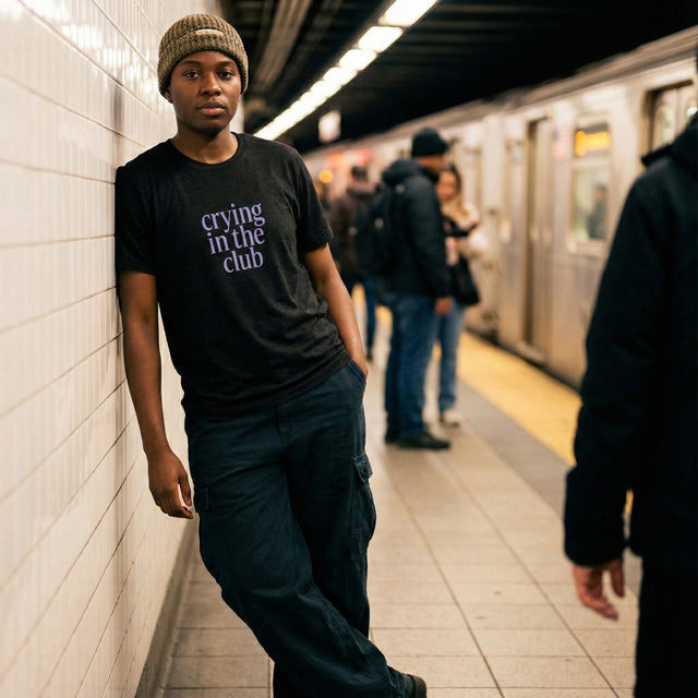 Unisex black heather t-shirt with lavender 'crying in the club' serif text, worn by a fashion model in an urban subway station setting.