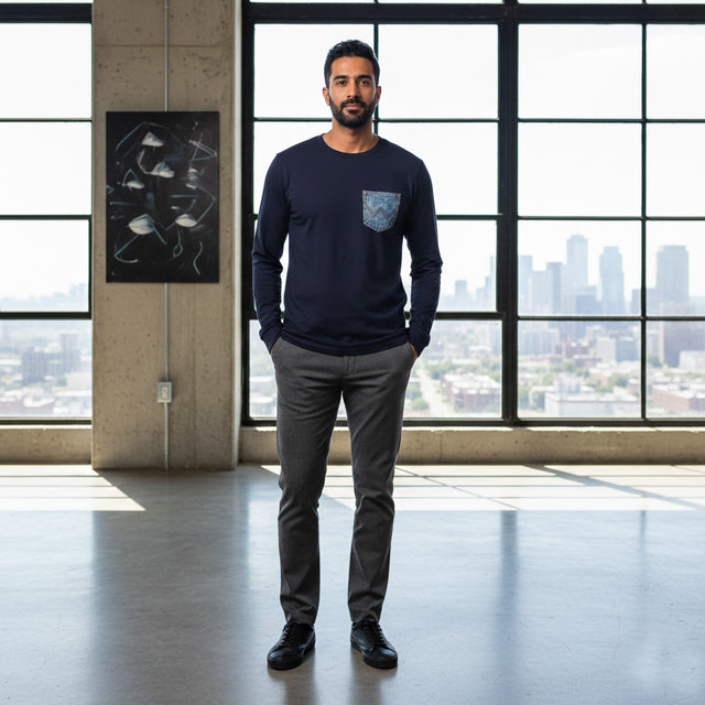 Man wearing a navy blue sweater with a pocket and gray pants standing in a modern room with large windows.