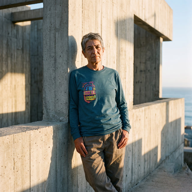 An older person stands outdoors against a concrete structure with the ocean in the background, wearing a teal long-sleeve shirt with a colorful graphic on the chest and brown pants.