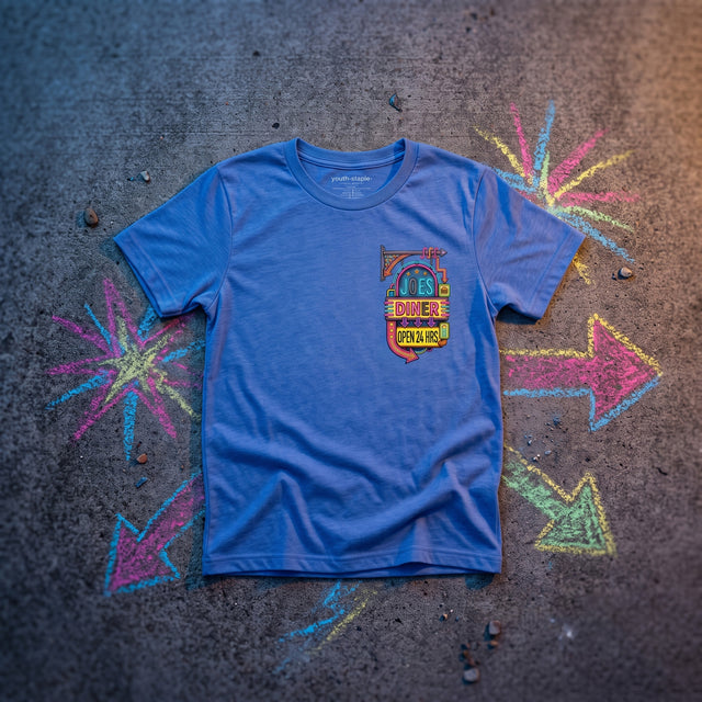 A blue short-sleeve graphic t-shirt laid flat on a dark textured ground with colorful chalk starburst and arrow drawings around it. The shirt features a small shield-style chest graphic with the words 'JESUS IS MY DINNER AND MY GOD'.