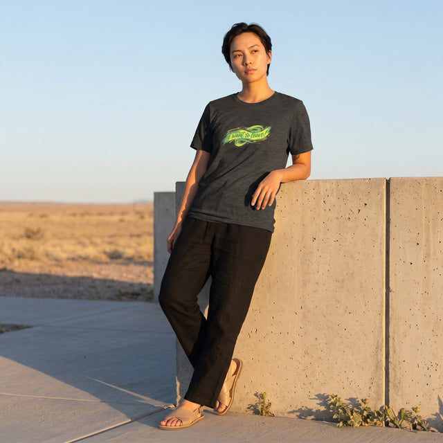 ロゴ入りの濃い色のTシャツを着た人物が、背景に砂漠の風景を背に、コンクリートの壁に寄りかかって立っている。