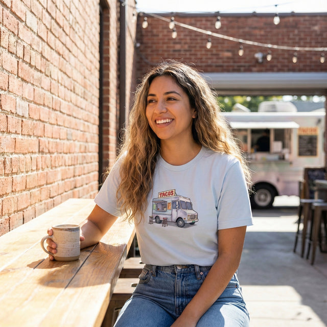 A lifestyle photograph of a young woman wearing the light blue ICHINICHI t-shirt with the watercolor taco truck graphic, smiling while holding a coffee cup on a sunny cafe patio.