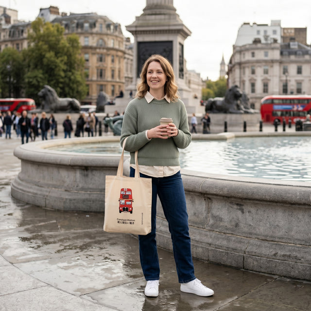 Woman holding a coffee cup and a tote bag with a red double-decker bus design, standing in front of Trafalgar Square.