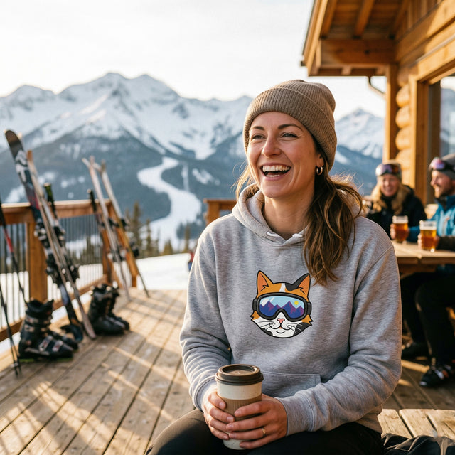 Woman in a gray hoodie with a cat graphic, holding a coffee cup, on a wooden deck with skis and mountains in the background.
