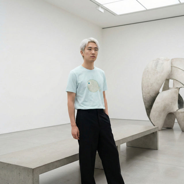 Ichinichi unisex staple t-shirt in heather ice blue featuring a minimalist pufferfish (fugu) illustration, worn by a male model in a modern art gallery setting, Japanese streetwear style.