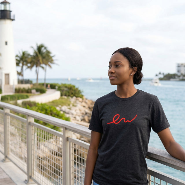 Woman wearing a black t-shirt with red text, standing by a waterfront with a lighthouse and palm trees in the background.