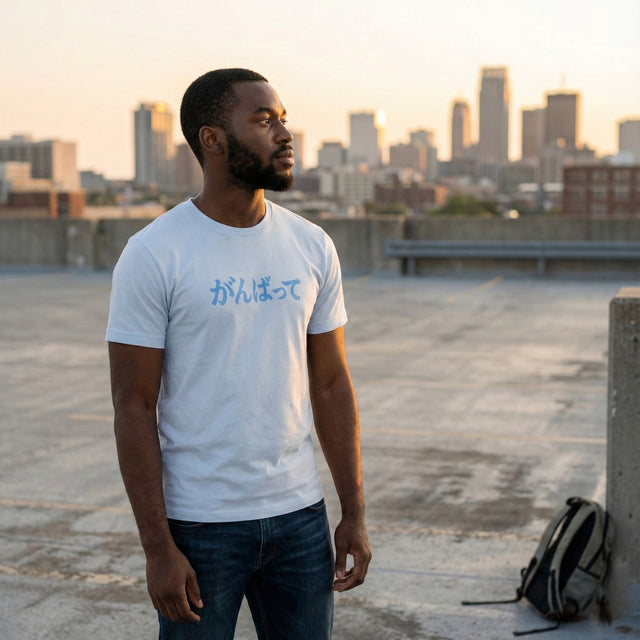 Young man standing on an urban rooftop wearing a light blue unisex t-shirt with blue Japanese text "Ganbatte" (Do Your Best) printed on the chest.