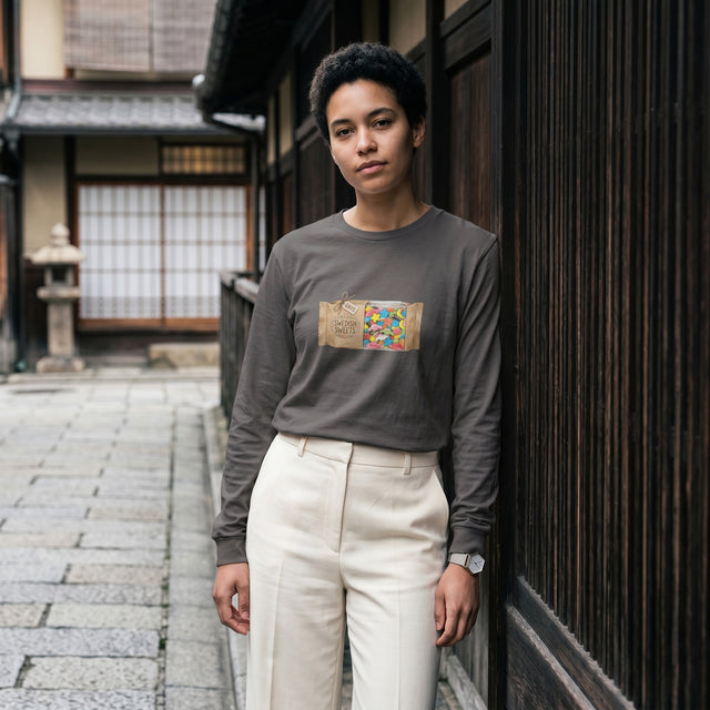 Person wearing a dark gray long-sleeve shirt with a colorful graphic and light-colored pants standing in front of traditional wooden architecture.