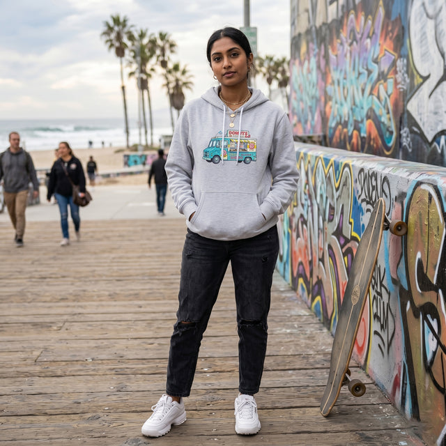 A streetwear-style candid portrait of a young South Asian woman, late 20s, wearing the light grey 'Daisy's Delicious Donuts' food truck hoodie on the Venice Beach Boardwalk in Los Angeles.