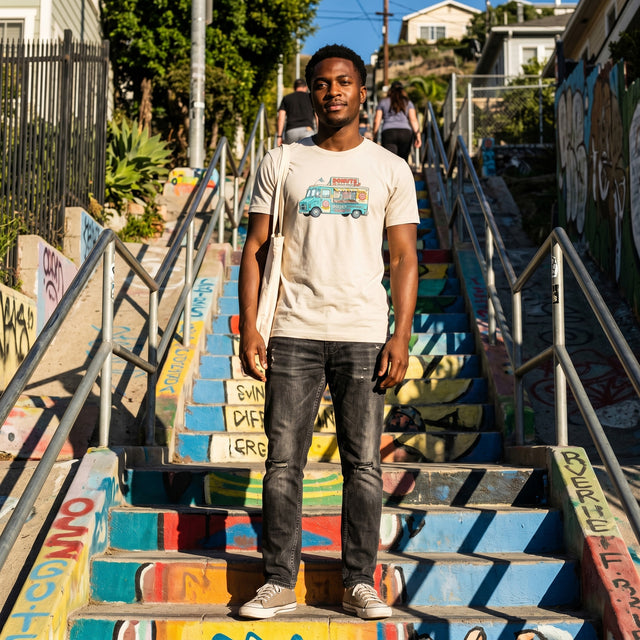 A young Nigerian-British man wearing the "Daisy's Delicious Donuts" heather dust t-shirt, standing on the colorful Silver Lake Stairs in Los Angeles.