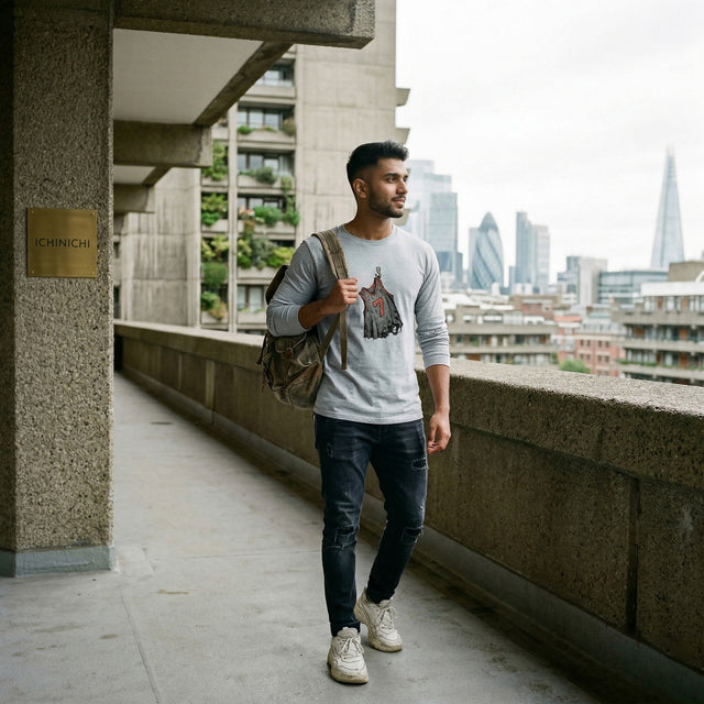 A high-quality lifestyle photograph of a 20s South Asian man wearing the 'Echoes of Effort' grey long-sleeve tee from image_24.png. The detailed graphic of the tattered jersey number 7 is sharply in focus against the textured concrete walls and background architecture of the Barbican Estate in London under a soft, overcast sky.