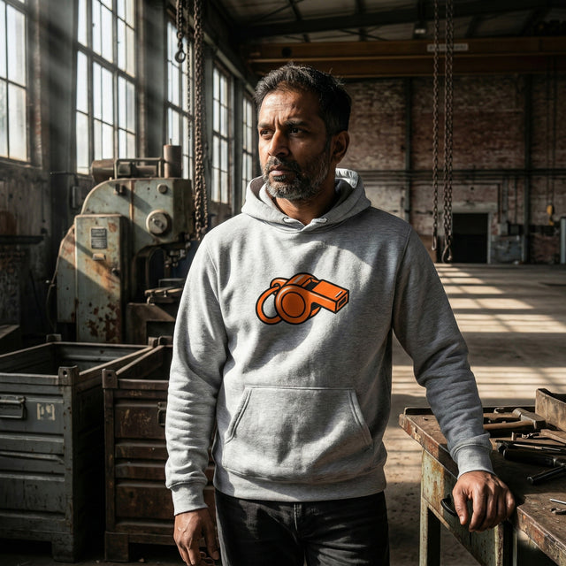 A middle-aged South Asian man wearing a grey athletic heather pullover hoodie with an orange whistle graphic, standing in a sunlit industrial hangar with a rugged, stoic expression.